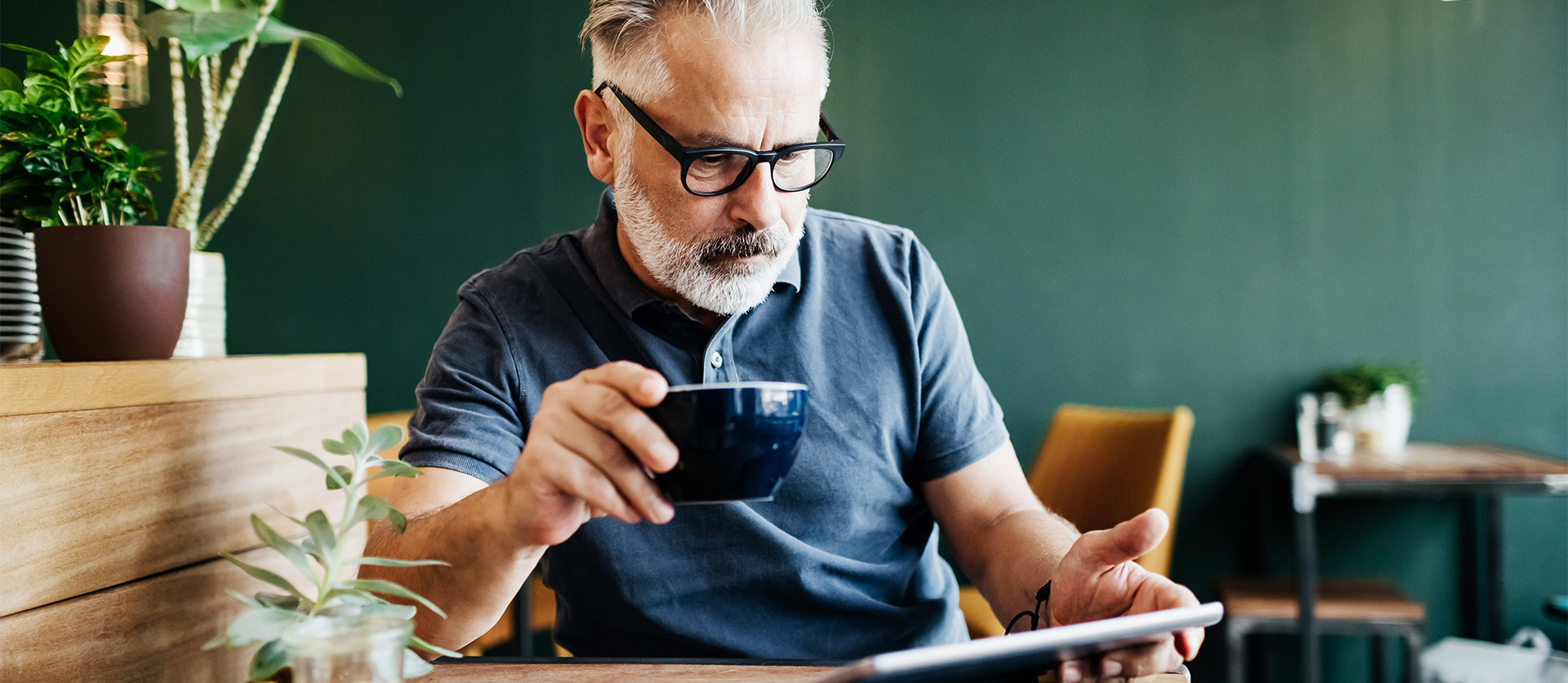 Man kijkt aandachtig naar zijn tablet met een kop koffie in zijn hand